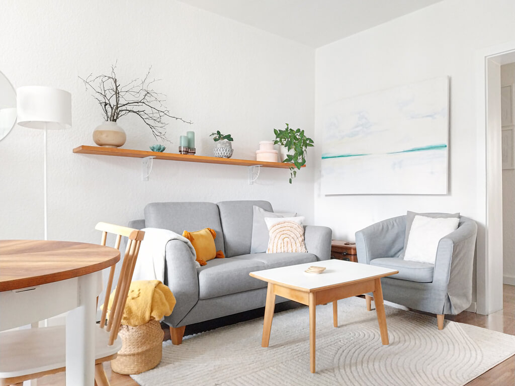 Skandi-Boho-Vintage Wohnzimmer mit grauem Sofa, hellem Holz, orangefarbenem Akzentkissen und hellen Wänden.